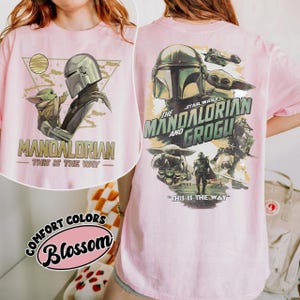 May include: Pink t-shirt featuring a graphic of The Mandalorian and Grogu from Star Wars. The front design includes the Mandalorian and Grogu with the text "MANDALORIAN - THIS IS THE WAY". The back design features the text "STAR WARS THE MANDALORIAN AND GROGU".