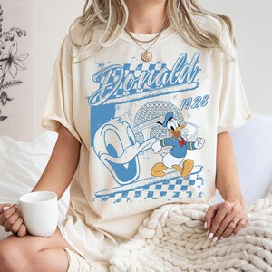 May include: Cream-colored t-shirt featuring a blue graphic of Donald Duck with the text "Donald 1928". The design includes a checkered pattern and an image of a building. The shirt has short sleeves.