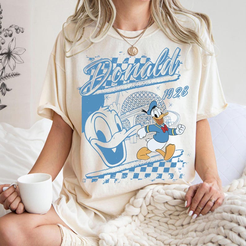 May include: Cream-colored t-shirt featuring a blue graphic of Donald Duck with the text "Donald 1928". The design includes a checkered pattern and an image of a building. The shirt has short sleeves.