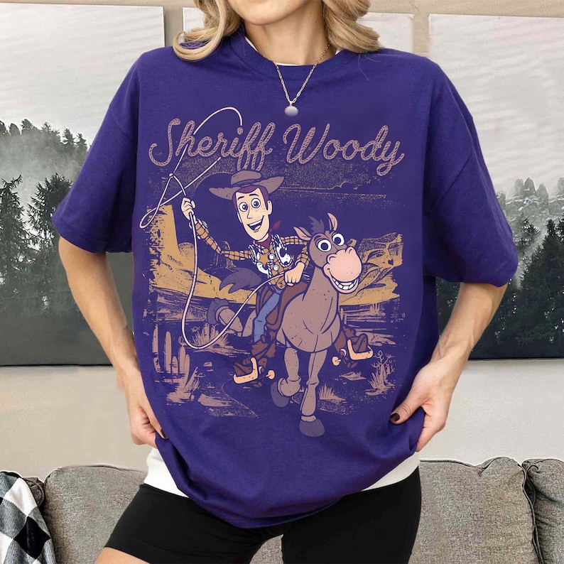 May include: A purple t-shirt featuring a graphic of Sheriff Woody riding a horse with the text "Sheriff Woody" in a stylized font. The design has a vintage, distressed look, with a desert landscape background.