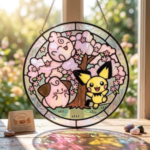 May include: A circular stained glass suncatcher featuring a yellow character with black-tipped ears and two pink creatures positioned beneath a tree with light pink blossoms. The piece is framed in black metal and hangs from a silver chain.