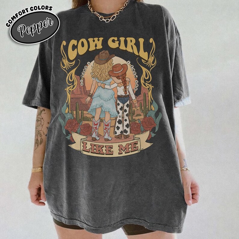 May include: A charcoal gray short-sleeve t-shirt featuring a graphic print of two figures in cowboy hats standing in a desert landscape. The text reads COW GIRL and LIKE ME. The design includes cacti, red roses, and a decorative banner frame.