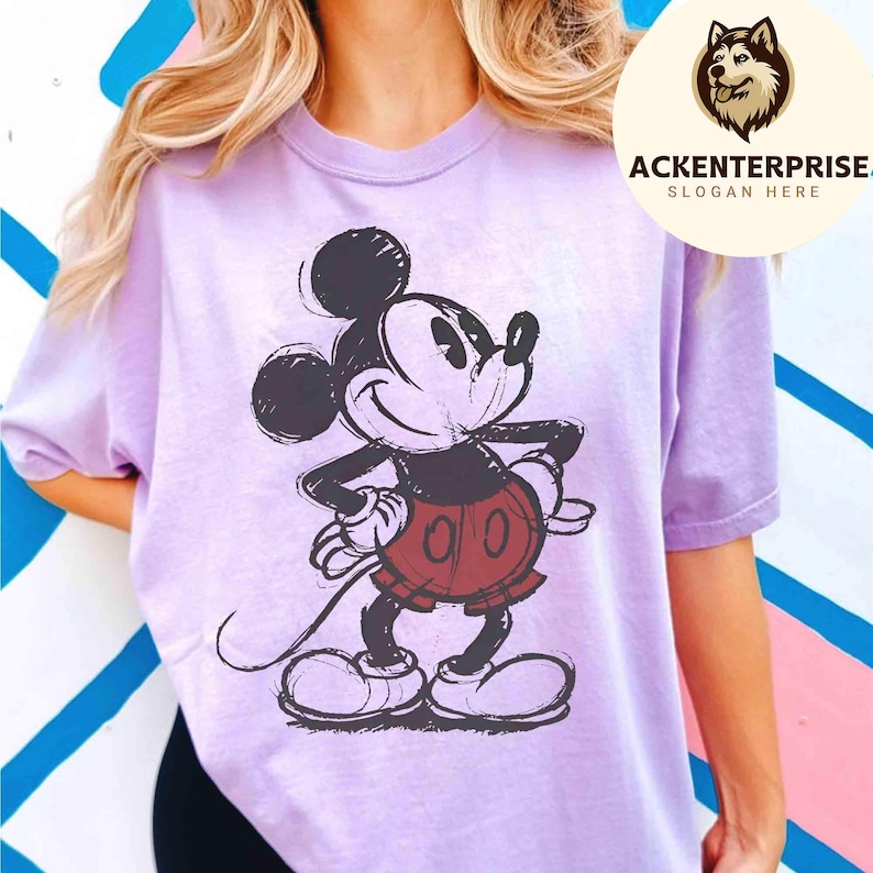 Puede incluir: Una camiseta de color lila claro con una imagen esbozada de Mickey Mouse en negro, rojo y blanco. La camiseta tiene mangas cortas y cuello redondo. La imagen incluye el texto "ACKENTERPRISE. SLOGAN HERE" en un logotipo circular.