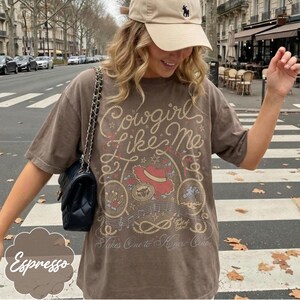 Puede incluir: Una camiseta marr&oacute;n de manga corta con un estampado gr&aacute;fico que incluye el texto Cowgirls Like Me y Takes One to Know One. El dise&ntilde;o presenta un sombrero de vaquero rojo, un lazo, notas musicales, estrellas y una herradura.