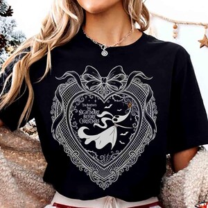 May include: Black t-shirt featuring a white graphic from "The Nightmare Before Christmas." The design includes a heart-shaped frame with a bow, the film's title, and a silhouette of Zero. Bats and spiderwebs complete the spooky theme.