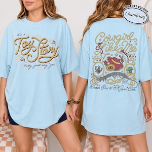 May include: A light blue t-shirt featuring a front graphic with rope-style text reading "it's a Toy Story Baby, just say 'yes'" and a back graphic with rope borders, a cowboy hat, a horseshoe, and text reading "Cowgirl like Me Takes One to Know One."