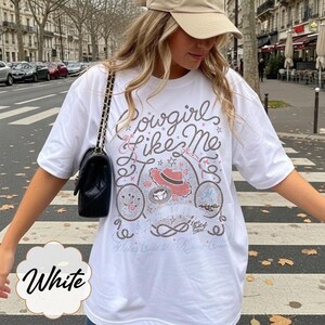 Puede incluir: Una camiseta blanca de manga corta con un estampado gr&aacute;fico que incluye el texto Cowgirl Like Me, Eyes full of stars y Takes One to Know One. El dise&ntilde;o presenta ilustraciones de un sombrero de vaquero, una herradura, una cuerda y notas musicales.