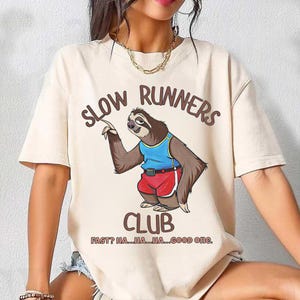 May include: Beige t-shirt with the text "SLOW RUNNERS CLUB" and a cartoon sloth wearing a blue tank top and red shorts. The sloth is pointing upwards. Below the club name is the text "FAST? HA...HA...HA...GOOD ONE."