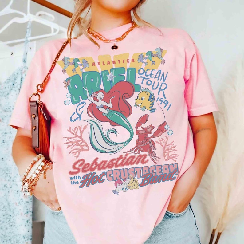 May include: Pink t-shirt featuring a graphic of Ariel the mermaid, Flounder, and Sebastian the crab. The design includes the text "Atlantica Ocean Tour 1991" and "Sebastian with the Hot Crustacean Band."