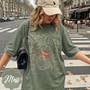 Puede incluir: Una camiseta de manga corta color verde oliva con un estampado gr&aacute;fico que incluye el texto Cowgirl like Me y Takes One to Know One. El dise&ntilde;o presenta un sombrero rojo, un lazo, notas musicales y estrellas. Se lleva un bolso negro acolchado.