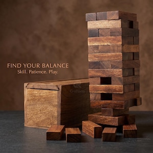Könnte beinhalten: Ein Holzklötzchen-Stapelspiel mit dem Text "FIND YOUR BALANCE Skill. Patience. Play." Das Spiel besteht aus rechteckigen Holzklötzen, von denen einige zu einem Turm gestapelt sind. Neben dem Turm befindet sich eine Holzkiste, mit verstreuten Klötzen.