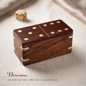 May include: A rectangular wooden box designed to resemble a domino, featuring a dark brown wood grain, inlaid brass dots on the top, and brass corner accents. A small, cream-coloured die with black pips sits nearby. Text reads: Dominoes HANDCRAFTED | PREMIUM ROSEWOOD.