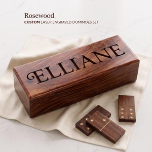 May include: A rectangular rosewood box with the name ELLIANE laser engraved on the lid, accompanied by three matching wooden dominoes featuring brass pins. The set is presented on a light-coloured fabric cloth, showcasing the natural wood grain.