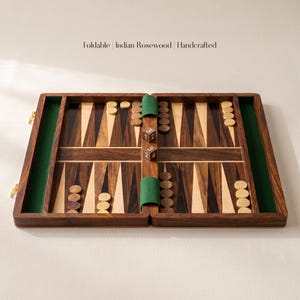 May include: A foldable Indian Rosewood handcrafted backgammon board featuring dark and light wood triangular points, green felt side rails, and circular game pieces. Two wooden dice with white numbers are positioned in the center of the board.