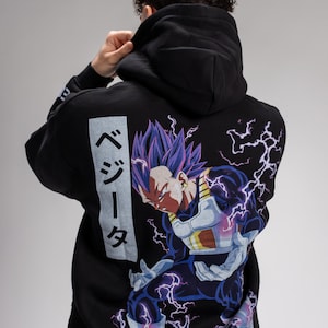 Op de afbeelding: Zwarte hoodie met een afbeelding van een personage met paars haar en bliksem. De hoodie heeft Japanse tekst aan de linkerkant. Het personage staat in een vechthouding. De hoodie is gemaakt van een zacht materiaal.
