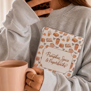 May include: A clear protective case for an e-reader featuring a repeating pattern of books, coffee cups, and bows. A central white rectangular label displays the text Pumpkin Spice & Paperbacks in a brown script font against a white background.