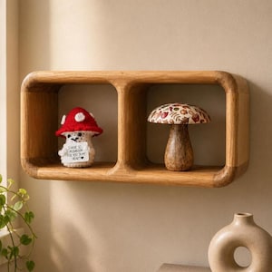 Puede incluir: Una estantería de madera con dos compartimentos cuadrados, cada uno con un objeto decorativo con temática de setas. Uno es una seta de ganchillo con gorro rojo y lunares blancos, sosteniendo un cartel. El otro es una seta de madera con gorro estampado.