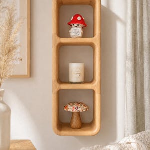 May include: A light oak-coloured, three-tiered shelf with rounded corners, mounted on a white wall. It displays a red-capped mushroom figurine, a candle, and a decorative mushroom. The shelf is a decorative item for a home.