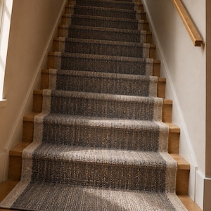 May include: A staircase with a runner rug. The runner is a dark grey with a lighter border, covering the centre of the wooden steps. A wooden handrail runs along the wall. A window is visible on the left side of the image.
