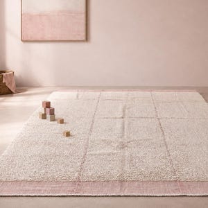May include: A rectangular area rug featuring a textured cream-colored center with a grid pattern and solid dusty pink borders on the top and bottom edges. The rug is placed on a smooth floor in a room with soft pink walls and various children's toys.
