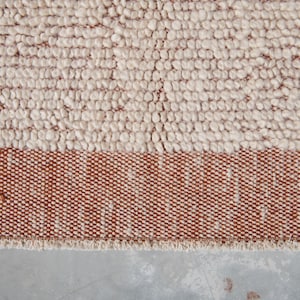 K&ouml;nnte beinhalten: Nahaufnahme eines strukturierten Teppichs mit zweifarbigem Design. Der obere Bereich weist eine cremefarbene, knotige Textur auf, w&auml;hrend der untere Bereich ein Band aus dicht gewebtem, rotbraunem Stoff aufweist. Der Teppich hat einen Fransenrand.