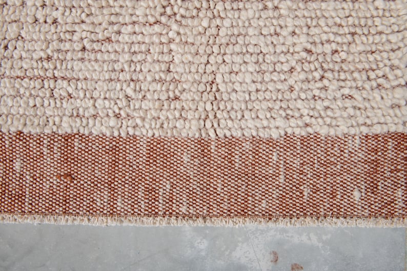 K&ouml;nnte beinhalten: Nahaufnahme eines strukturierten Teppichs mit zweifarbigem Design. Der obere Bereich weist eine cremefarbene, knotige Textur auf, w&auml;hrend der untere Bereich ein Band aus dicht gewebtem, rotbraunem Stoff aufweist. Der Teppich hat einen Fransenrand.