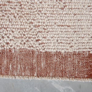 K&ouml;nnte beinhalten: Nahaufnahme eines strukturierten Teppichs mit zweifarbigem Design. Der obere Teil weist eine cremefarbene, noppige Textur auf, w&auml;hrend der untere Teil ein durchgehendes Band aus rotbraunem Webmaterial aufweist. Der Teppich hat einen Fransenrand.