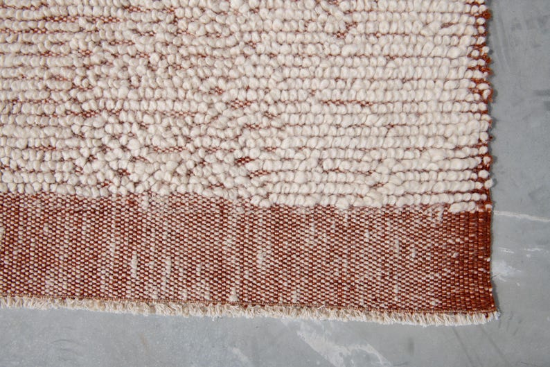 K&ouml;nnte beinhalten: Nahaufnahme eines strukturierten Teppichs mit zweifarbigem Design. Der obere Teil weist eine cremefarbene, noppige Textur auf, w&auml;hrend der untere Teil ein durchgehendes Band aus rotbraunem Webmaterial aufweist. Der Teppich hat einen Fransenrand.