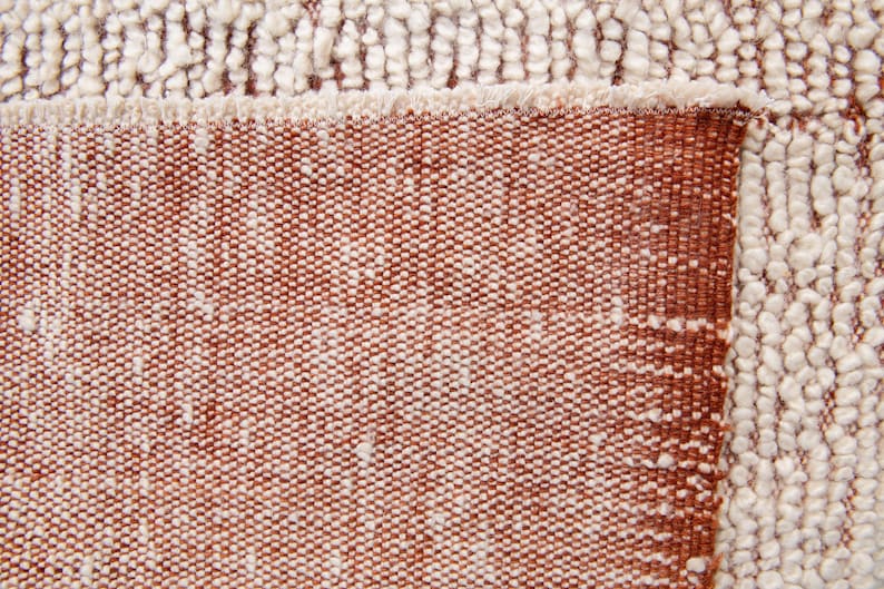 K&ouml;nnte beinhalten: Nahaufnahme eines strukturierten Teppichs in Braun- und Off-White-T&ouml;nen. Der Teppich hat ein gewebtes Design mit einem Fransenrand. Die Textur ist eine Kombination aus geschlungenen und flachen Webarten und erzeugt eine optisch ansprechende Oberfl&auml;che.