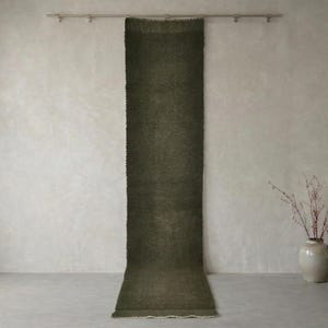 Könnte beinhalten: Ein langer, olivgrüner Teppich hängt an einer Metallstange vor einer neutralen Wand. Der Teppich hat eine strukturierte Optik und einen Fransenrand. Eine Vase mit Zweigen und eine Holzbank befinden sich im Hintergrund, was auf ein minimalistisches Wohndekor-Thema hindeutet.