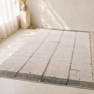 Pode incluir: Um tapete retangular branco sujo com uma borda e listras verticais cinza escuro. O tapete está colocado sobre um chão claro em uma sala bem iluminada. O design do tapete é simples e moderno.