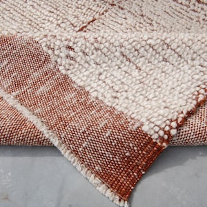 K&ouml;nnte beinhalten: Nahaufnahme eines strukturierten Teppichs mit einer gefalteten Ecke. Der Teppich weist ein gewebtes Design mit abwechselnden Abschnitten in Off-White- und Rotbraun-T&ouml;nen auf. Die Kanten sind mit einem Fransen versehen.