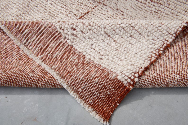 K&ouml;nnte beinhalten: Nahaufnahme eines strukturierten Teppichs mit einer gefalteten Ecke. Der Teppich weist ein gewebtes Design mit abwechselnden Abschnitten in Off-White- und Rotbraun-T&ouml;nen auf. Die Kanten sind mit einem Fransen versehen.