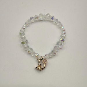 Op de afbeelding: Een armband van heldere kristallen kralen met een goudkleurige bedel. De bedel heeft de vorm van de letter "D" met een kleine roze bloem en groene bladeren. De armband ligt op een witte achtergrond.