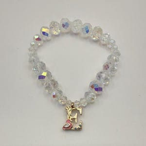 Puede incluir: Pulsera de cuentas transparentes con brillo iridiscente. La pulsera presenta un dije dorado con la letra "E" y un diseño de pájaro rosa. Las cuentas están facetadas, reflejando la luz.