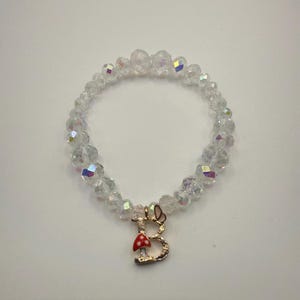 Puede incluir: Una pulsera de cuentas de cristal transparente con un dije dorado. El dije presenta un hongo rojo y blanco y la letra "B". La pulsera está hecha de pequeñas cuentas redondas e iridiscentes.