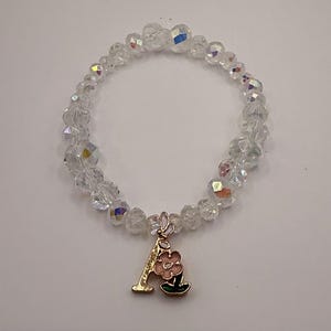 Puede incluir: Pulsera de cuentas de cristal transparente con un dije dorado. El dije tiene la forma de la letra "A" con una flor rosa y hojas verdes. La pulsera está sobre un fondo blanco.