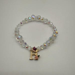 Op de afbeelding: Een heldere, iriserende kralenarmband met een goudkleurige bedel. De bedel heeft de letter "H", een kleine bloem en een paarse accent. De armband is gemaakt van facetkralen.
