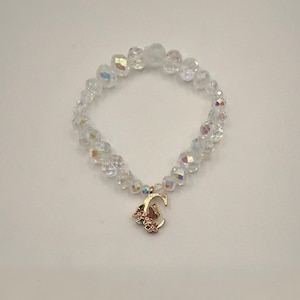 Op de afbeelding: Een heldere kralenarmband met een iriserende glans. De armband heeft een goudkleurige bedel in de vorm van de letter "C" met kleine roze bloemen. De kralen zijn facetgeslepen en reflecteren licht.