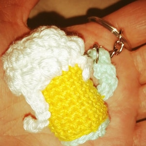 Peut inclure: Porte-clés en forme de chope de bière crochetée, avec une mousse blanche et un corps jaune. Le porte-clés est attaché à un anneau en métal argenté. La chope de bière est tenue dans une main.