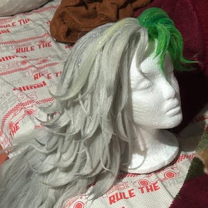 Può includere: Una parrucca grigia con una sezione verde sulla parte superiore, acconciata su una testa di polistirolo bianca. La parrucca ha capelli lunghi e ondulati con accenti glitterati. La testa poggia su un tessuto fantasia con la scritta "RACE THE RULE THE".