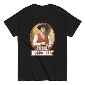 May include: Black t-shirt featuring a vintage-style graphic. The graphic is an oval portrait of a man in a hat and vest, with the text "I'M YOUR HUCKLEBERRY" in a red and white font. The design evokes a classic Western theme.