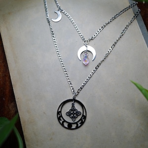 May include: A layered necklace with silver chains and pendants. The top chain features a crescent moon charm, while the middle chain has a crescent moon with a crystal pendant. The bottom chain has a black circular pendant with a Celtic knot design.