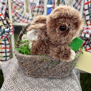 May include: A plush brown bunny with white accents sits in a woven basket with faux green grass. The basket is decorated with silver glitter and rests on a gray patterned pillow. A small green tag is attached.
