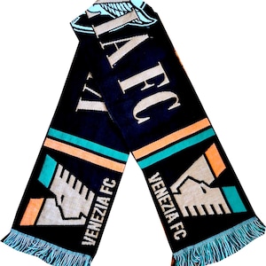 May include: A black knit scarf featuring the text VENEZIA FC in white lettering. The design includes a stylized V logo in white, orange, and teal, along with horizontal stripes in orange and teal. The ends of the scarf are finished with teal fringe.