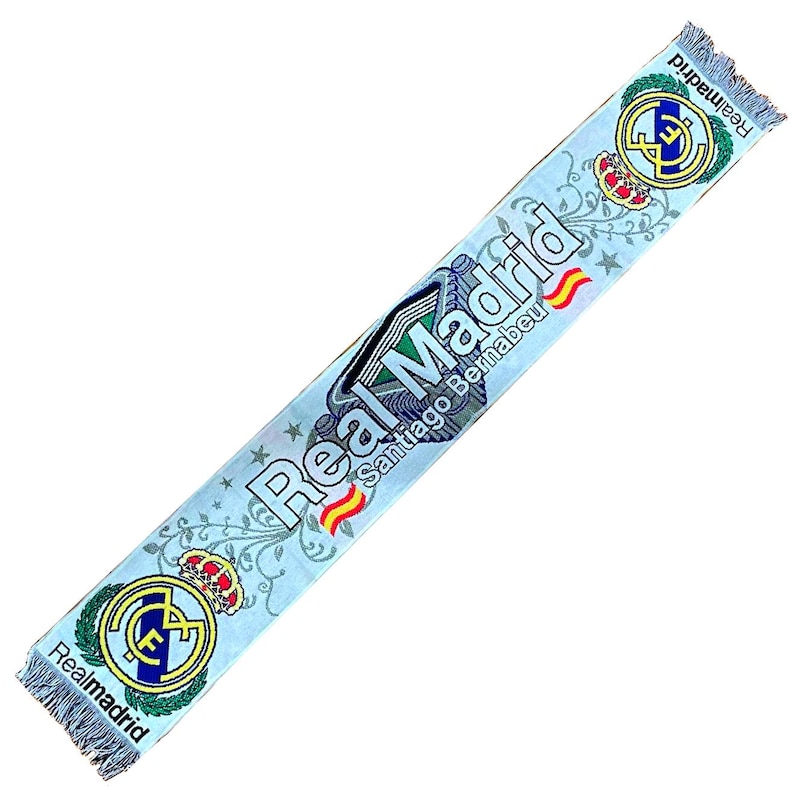 May include: Light blue Real Madrid scarf with the team's logo and the words "Real Madrid Santiago Bernabeu" printed on it. The scarf has a fringed edge and a repeating floral pattern.