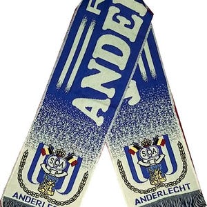 Puede incluir: Bufanda azul y blanca con la palabra "ANDERLECHT" en letras blancas grandes. La bufanda presenta un emblema del equipo con una corona, banderas y las letras "SCA". La bufanda tiene un borde con flecos.