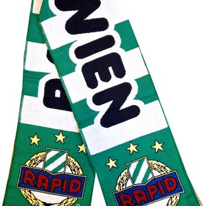 Könnte beinhalten: Grün-weißer Schal mit dem Wort "WIEN" in großen schwarzen Buchstaben. Der Schal zeigt das Wort "RAPID" in einem rot-blauen Emblem, umgeben von goldenen Sternen und einem grün-weißen Streifendesign. Der Schal hat einen Fransenrand.