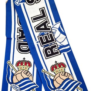 May include: A blue and white knitted scarf with the word "REAL" in black block letters. The scarf features a design of a soccer ball with a crown and a flag. The scarf has a fringe at the bottom.