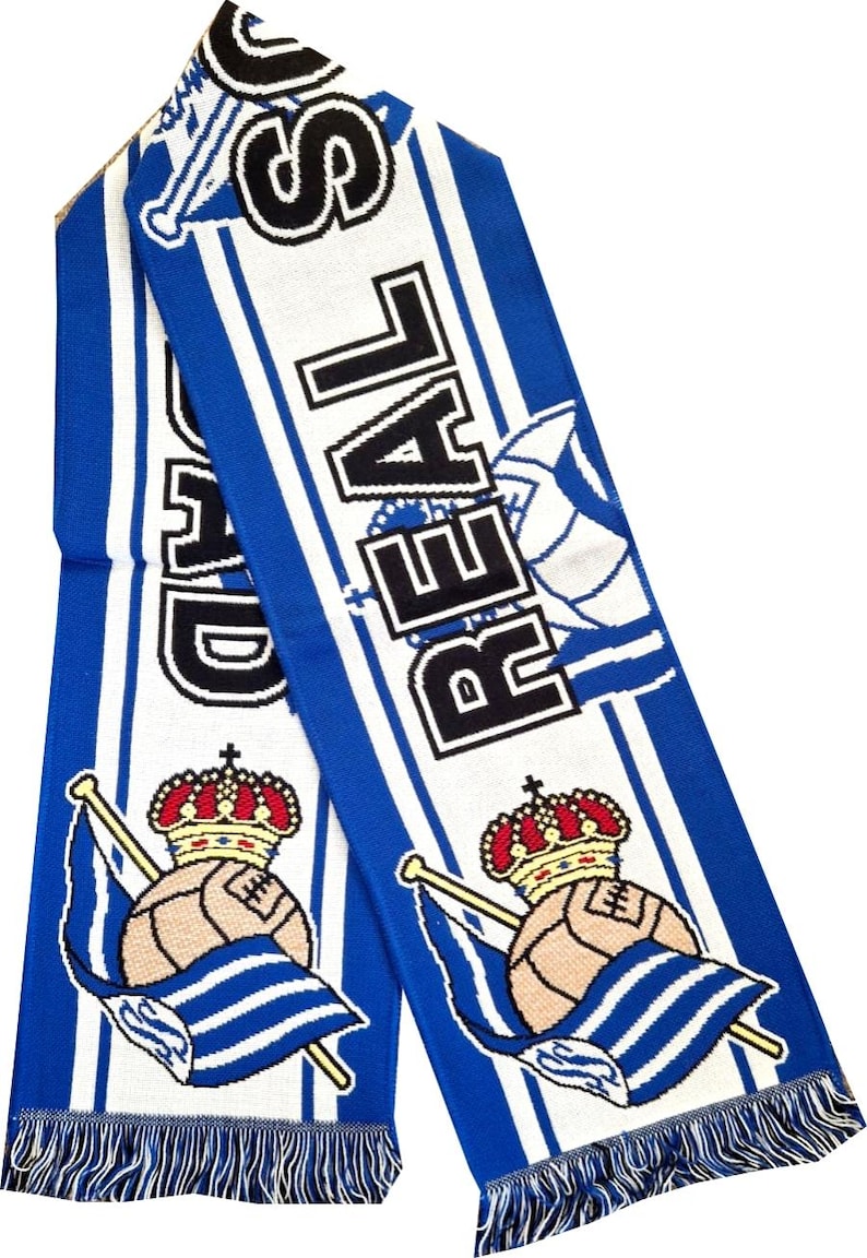May include: A blue and white knitted scarf with the word "REAL" in black block letters. The scarf features a design of a soccer ball with a crown and a flag. The scarf has a fringe at the bottom.